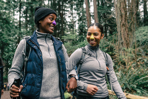 Two people smiling candidly together while hiking in the woods. They have bright colored, reef safe, vegan sunscreen on their noses. 